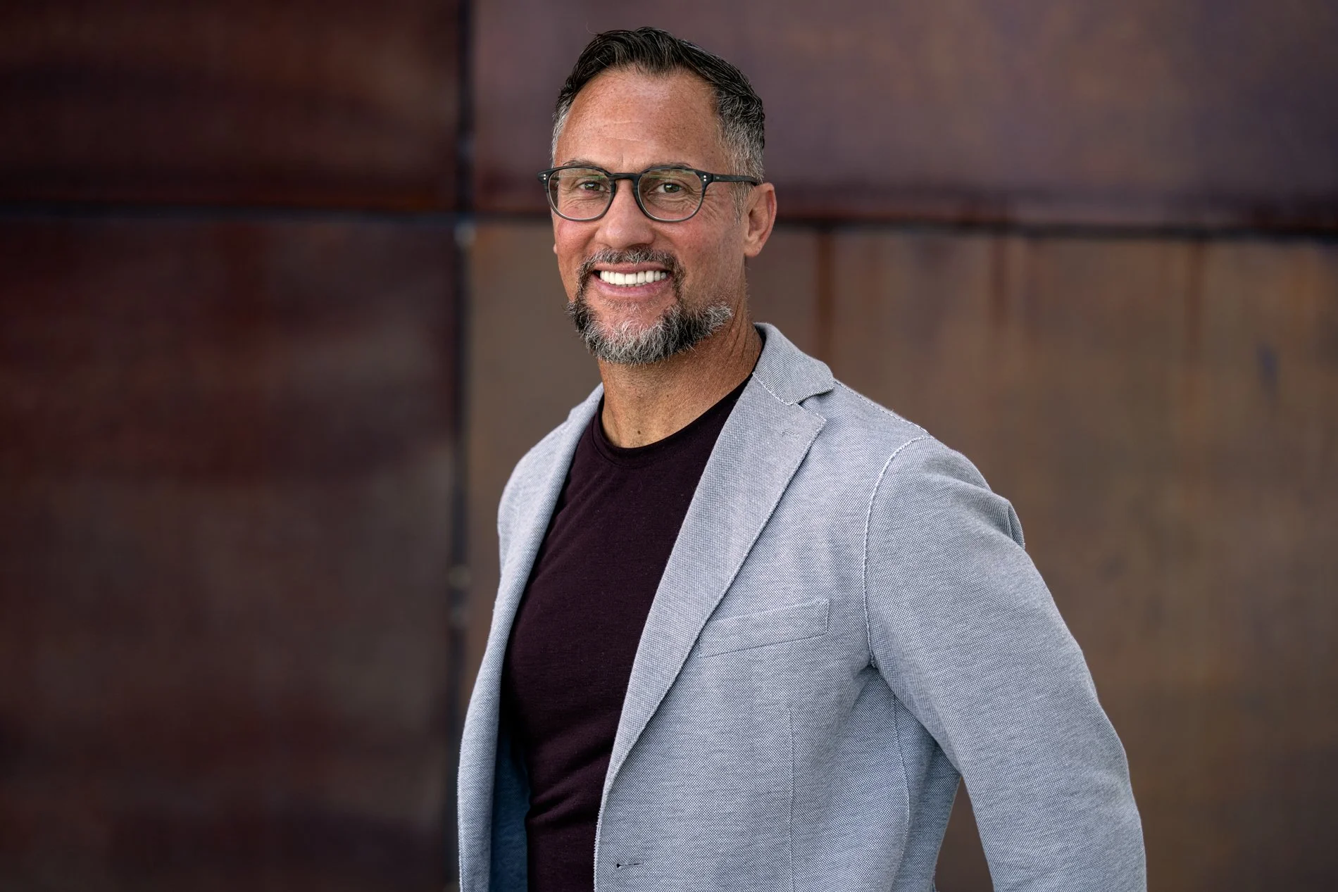 A middle-aged man with glasses, a beard and mustache, smiling, wearing a light gray blazer and a black shirt, standing outdoors against a dark brown, textured wall.