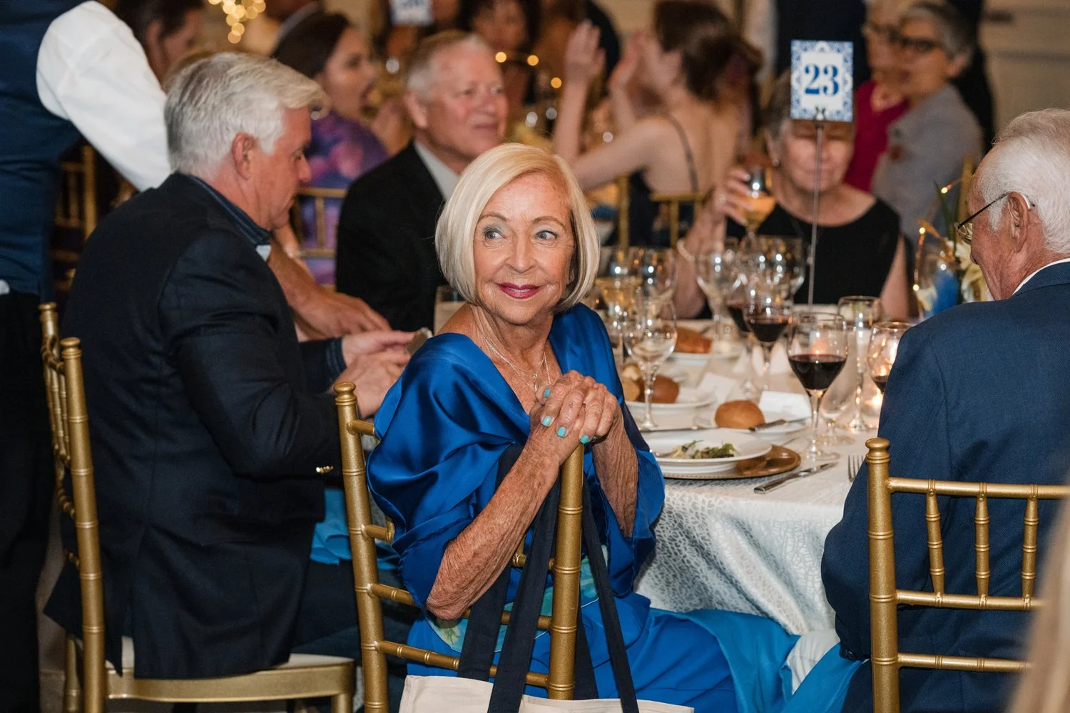 An elderly woman in a blue dress sitting at a banquet table during a formal event, surrounded by people. She has short, white hair and is smiling, with her hands clasped together on the table. The table is set with wine glasses, bread rolls, and plat