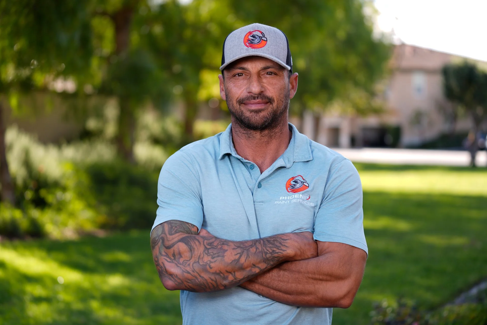 A man with a beard and tattoos on his arms stands outdoors, crossing his arms, wearing a light blue polo shirt and a gray baseball cap with a Phoenix Paint Services logo. Trees and a house are in the background.