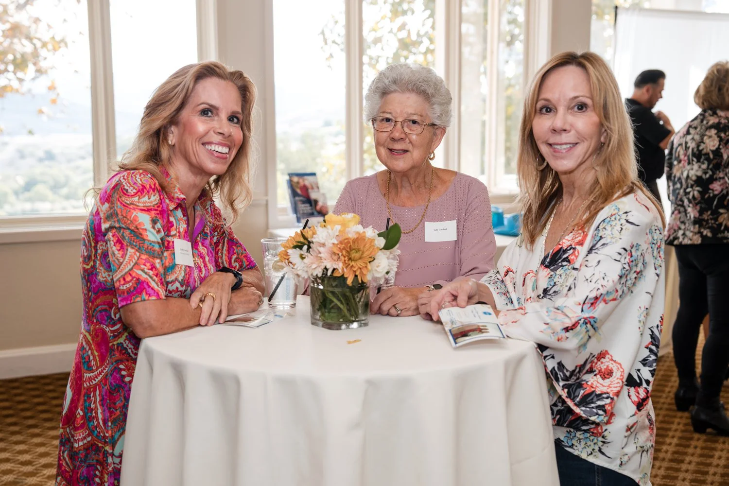 Three women sitting at a round table with a vase of flowers, smiling at a gathering or event, with windows in the background.