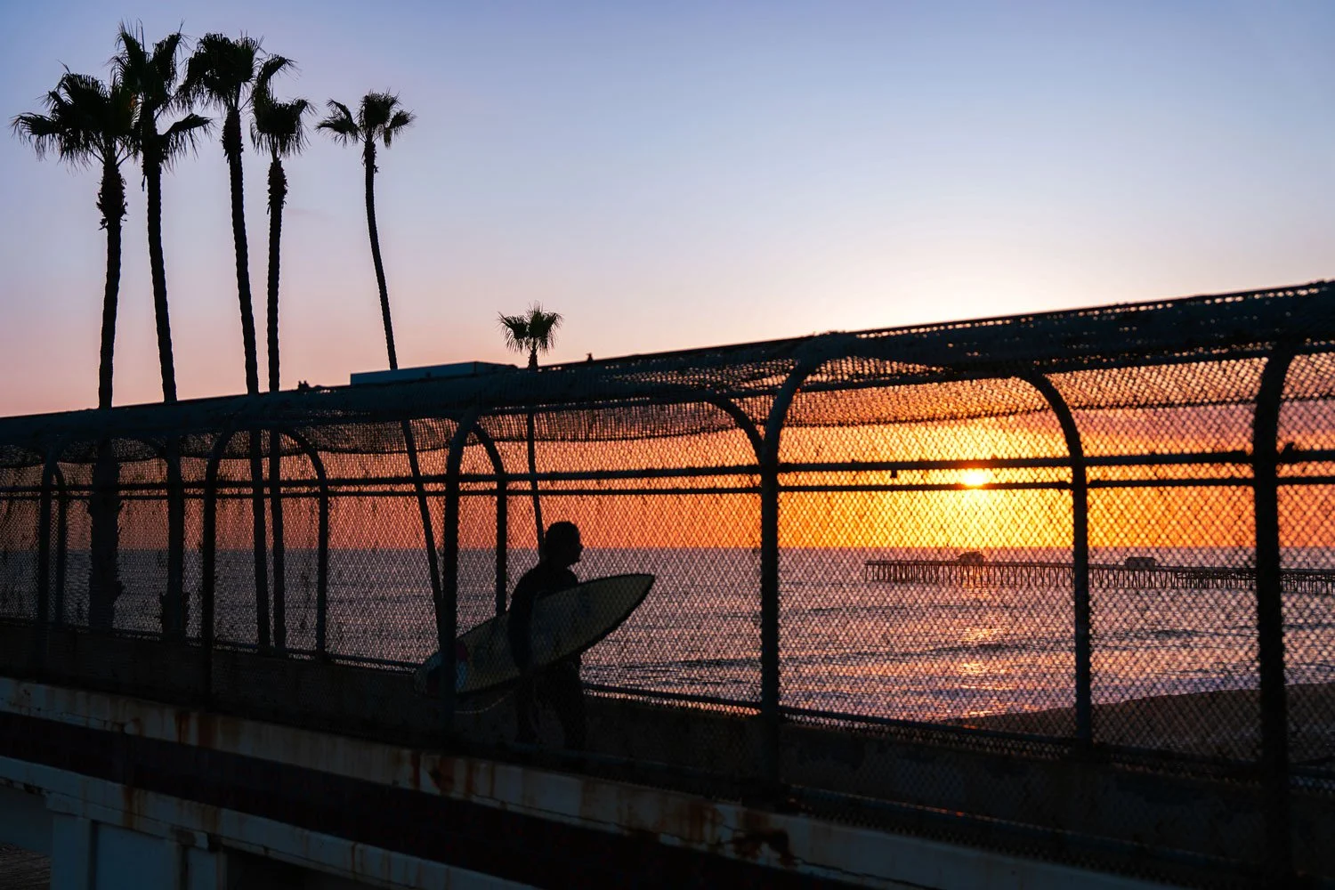 Silhouette of a person carrying a surfboard walking along a fenced pier at sunset, with palm trees in the background and the ocean extending toward a pier in the distance.