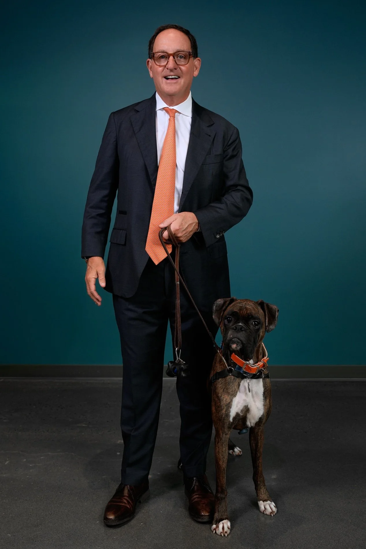 A man in a dark suit, white shirt, and orange tie standing with a brown and black brindle boxer dog on a leash against a teal background.