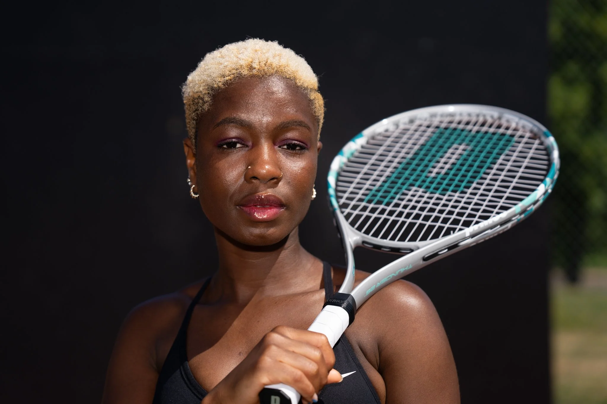 A woman with short, blonde, curly hair holding a tennis racket on a tennis court. She has earrings, a nose piercing, and is wearing a black sports top.