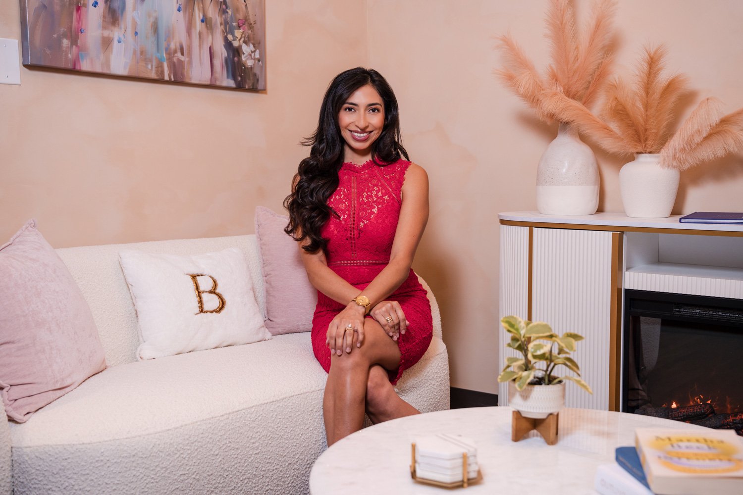 A woman with long dark hair sitting on a light-colored couch in a cozy, modern living room, wearing a red lace dress, smiling at the camera. Decor includes pillows, a painting, a white fireplace with pampas grass in vases, a small potted plant, and a