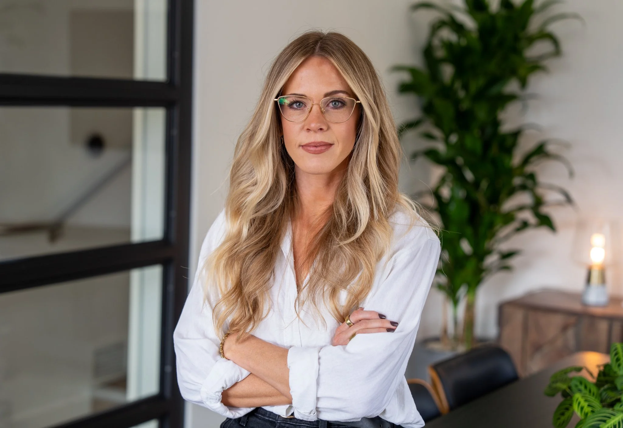 A woman with long blonde hair, wearing glasses and a white blouse, standing indoors with her arms crossed and looking at the camera. There is a large green plant in the background.