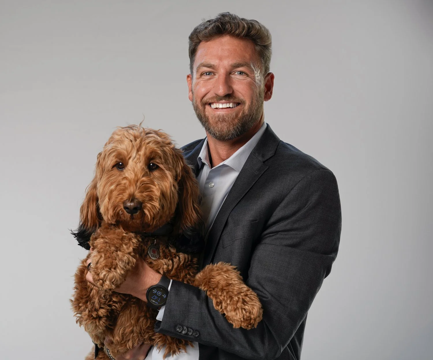 A man in a dark suit holding a brown curly-haired dog, both smiling at the camera against a plain background.