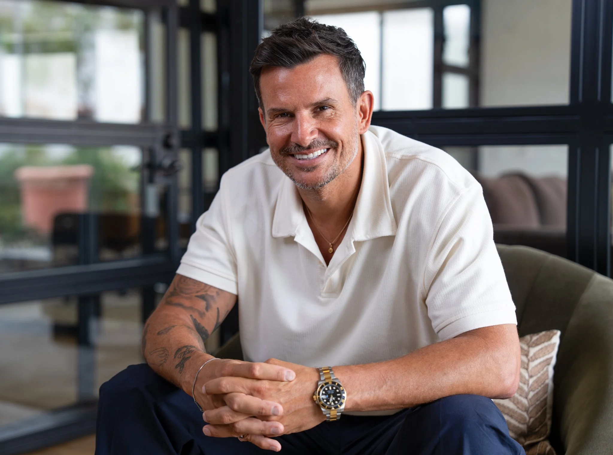 A smiling man with dark hair, wearing a white shirt, sitting indoors in front of a glass wall, with a potted plant outside. He is accessorized with a wristwatch and a gold necklace.