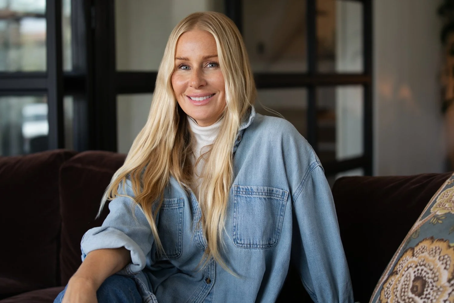 A woman with long blonde hair smiling, wearing a light denim jacket over a white turtleneck, sitting on a dark brown couch in a modern indoor setting.