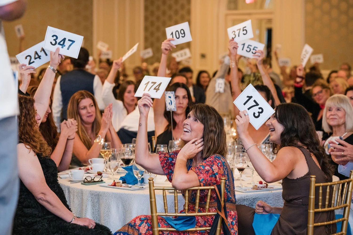 People seated at tables in a banquet hall, holding up number placards, likely for an auction or contest.