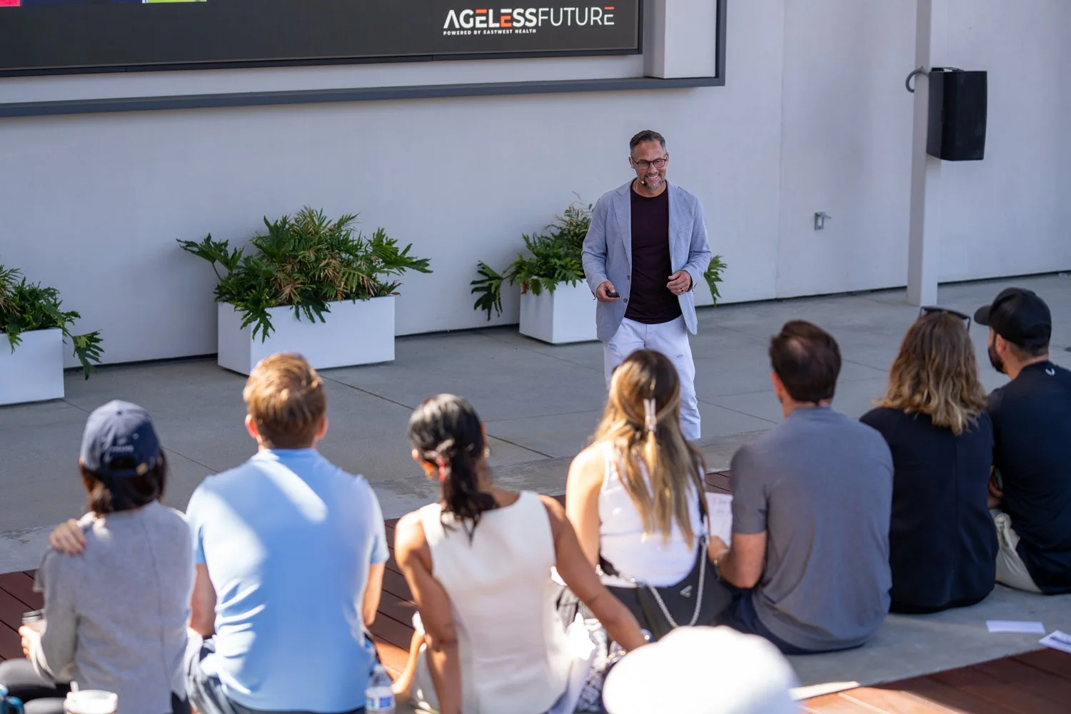 A man giving a presentation outdoors to a seated group of people with a sign reading "Ageless Future" in the background.
