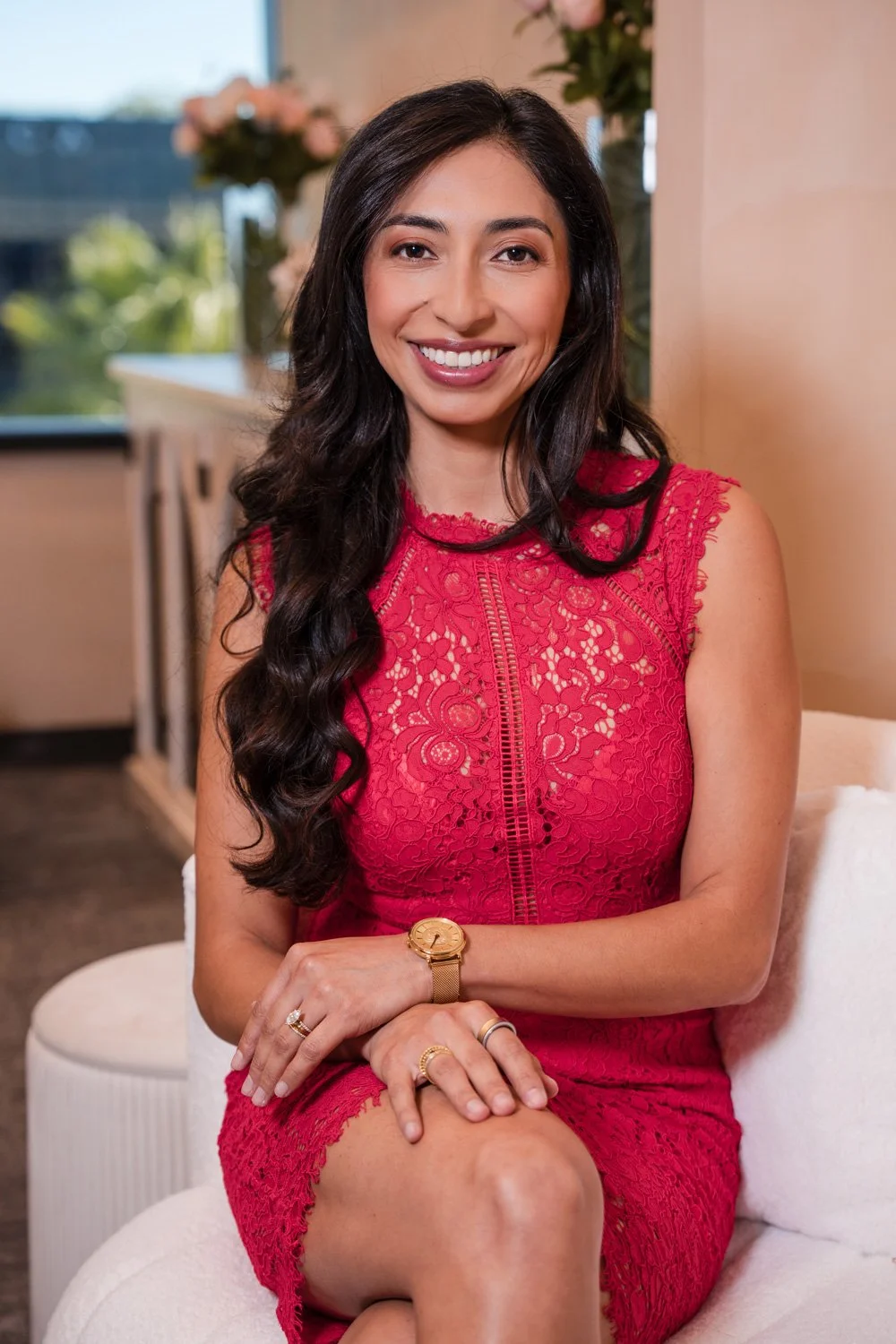 A woman with long dark hair and a bright smile sitting on a white chair in a well-lit room, wearing a red lace dress and gold watch.