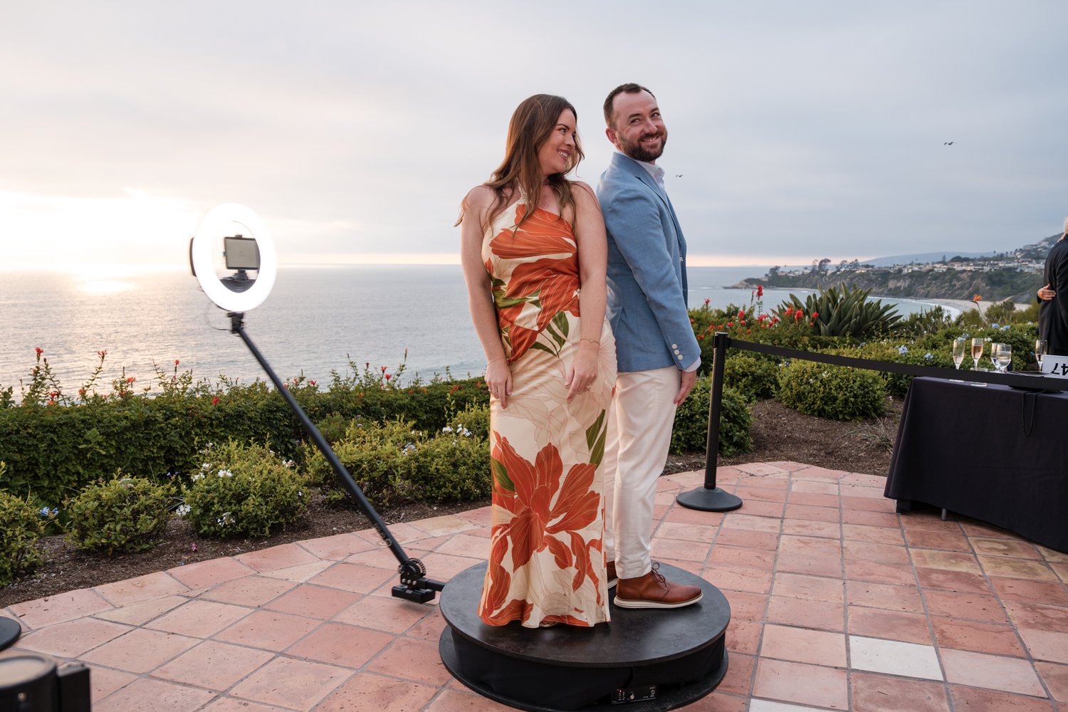 A woman and a man stand together on a circular platform outdoors near a coastline during sunset, with a ring light and camera setup nearby. The woman wears a floral dress with orange and green patterns, and the man wears a blue blazer, white pants, a
