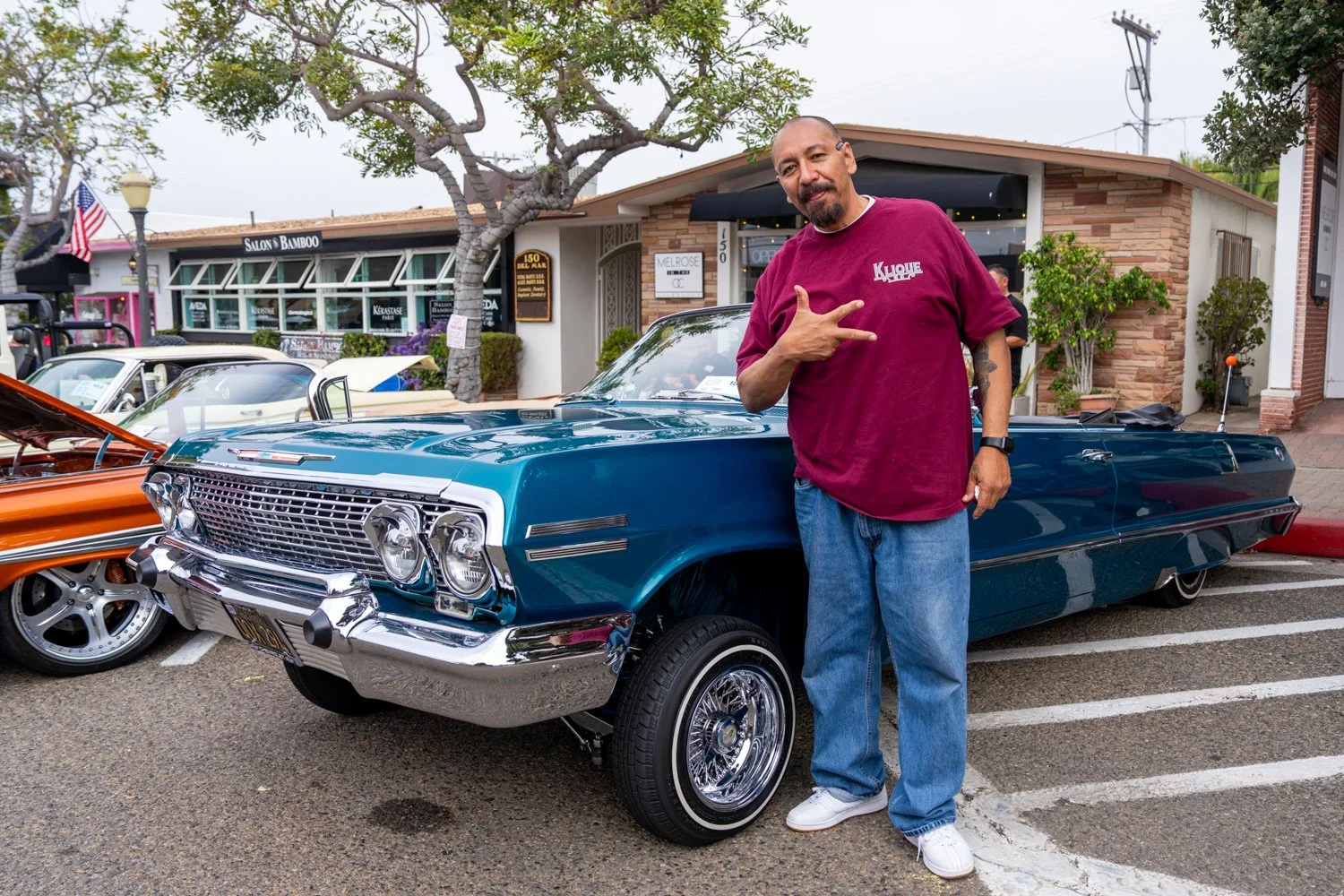 Man standing next to a classic blue car at a car show, pointing to his shirt with the word 'Kulture' on it, in front of a building with a sign reading 'Salon Bamboo.'