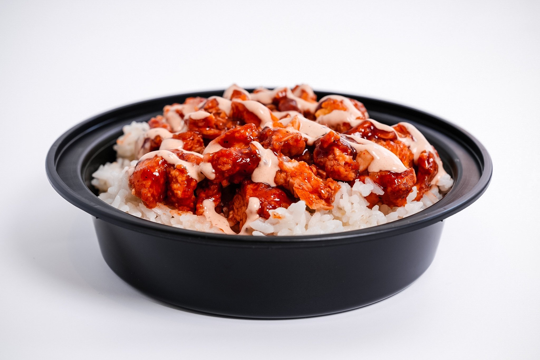 A black round container filled with white rice topped with saucy, spicy-looking pieces of chicken and drizzled with a creamy white sauce, set against a plain white background.