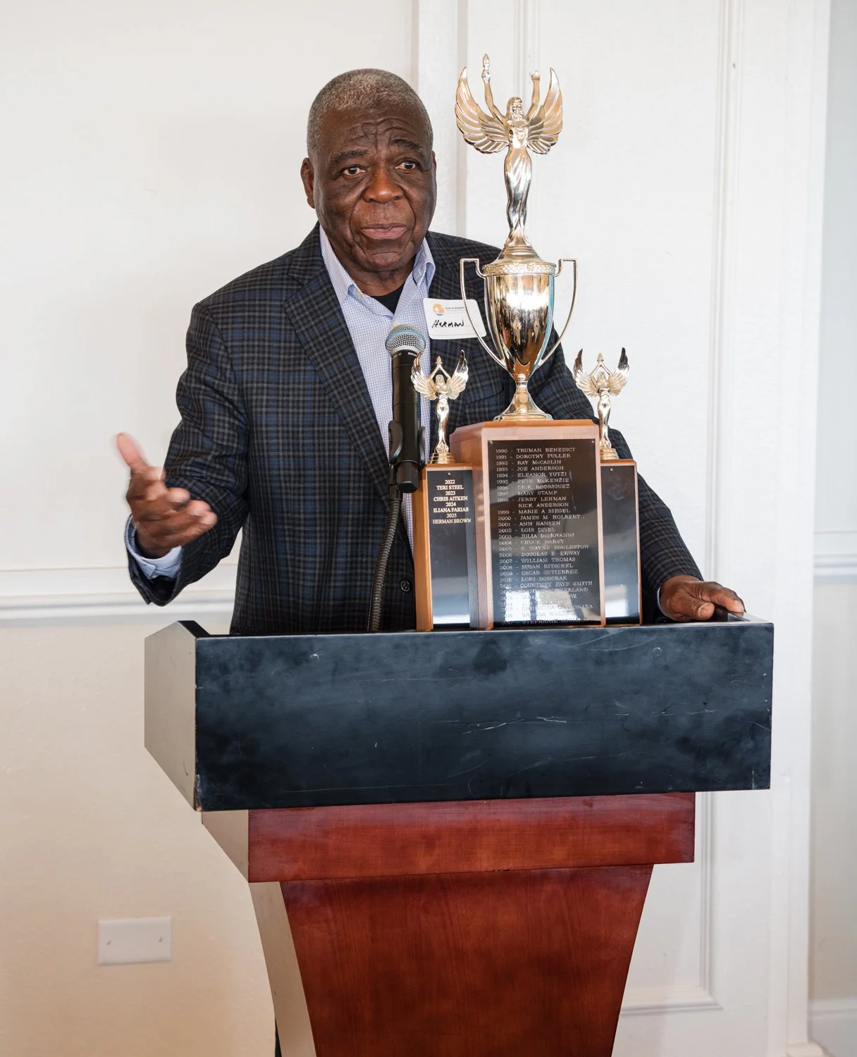 An elderly man in a suit standing at a podium with a microphone, holding a large trophy with a plaque, during a ceremony.