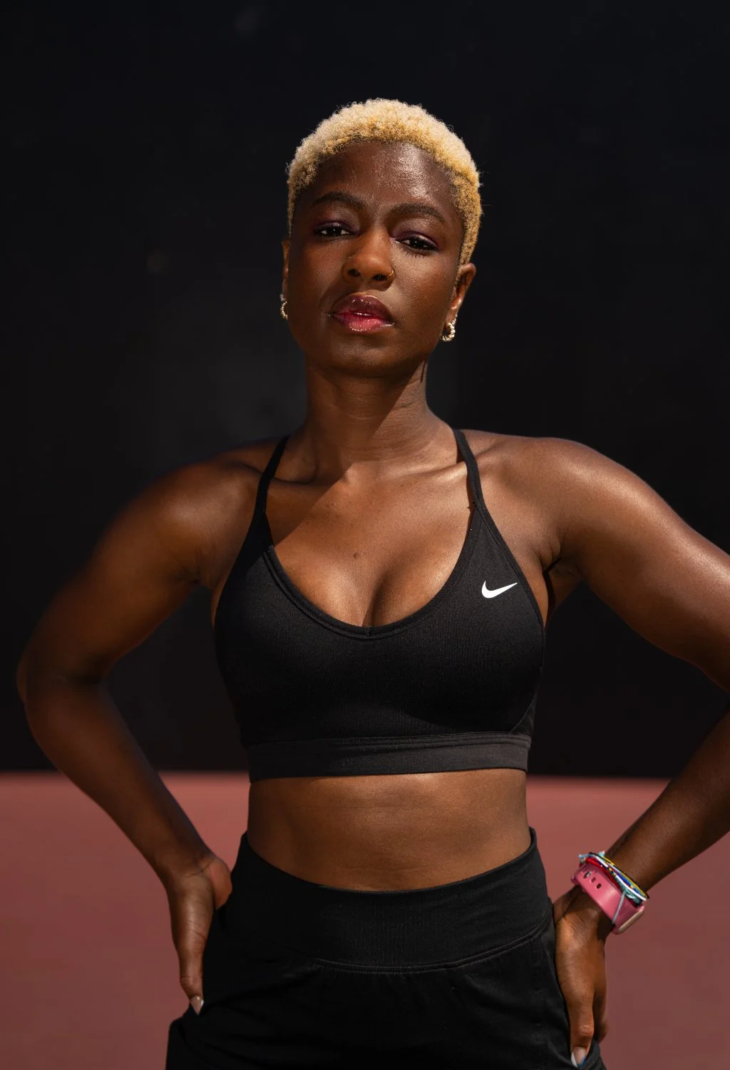 A confident woman with short blonde hair, wearing a black Nike sports bra and black athletic pants, standing with hands on hips against a dark background.