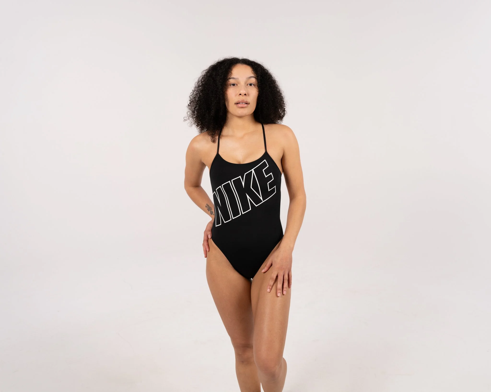 A woman with curly black hair and a tattoo on her right arm wearing a black Nike swimsuit standing against a plain light gray background.
