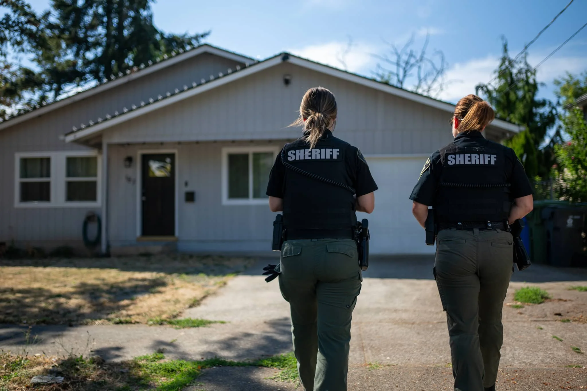 Two female sheriff deputies with back patches labeled 'SHERIFF' walking on a sidewalk in residential neighborhood.