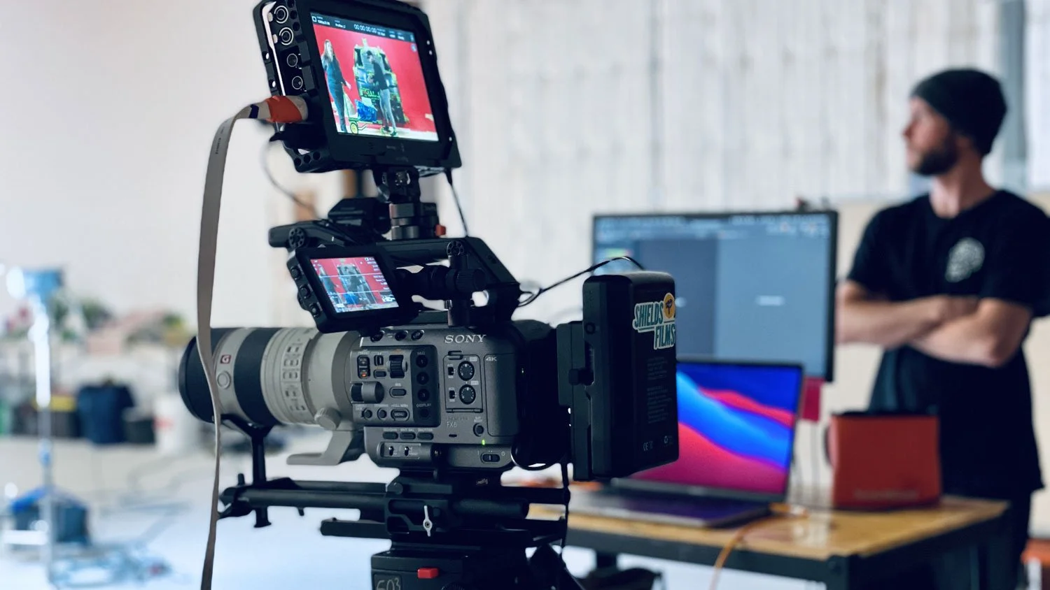 Professional camera recording a video of a man standing in front of a computer setup during a video shoot.