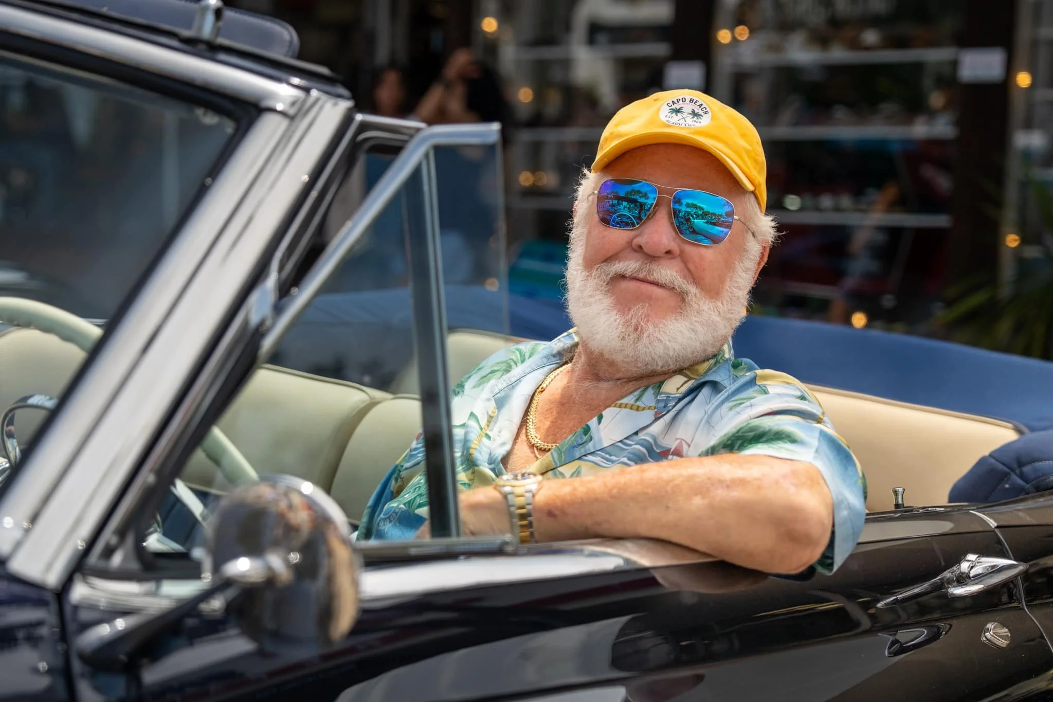 An older man with white hair and a beard driving a classic black convertible car, wearing sunglasses and a yellow cap with a tropical logo, and a colorful Hawaiian shirt.