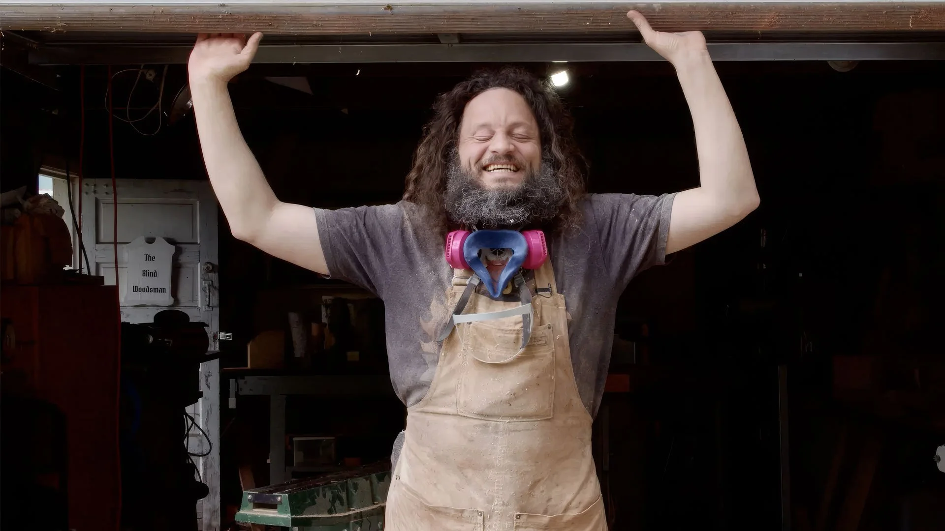 A man with a beard and curly hair, smiling with eyes closed, raising his arms holding a wooden beam. He is wearing a gray t-shirt, a tan apron, and pink protective goggles around his neck, standing in a woodworking workshop.