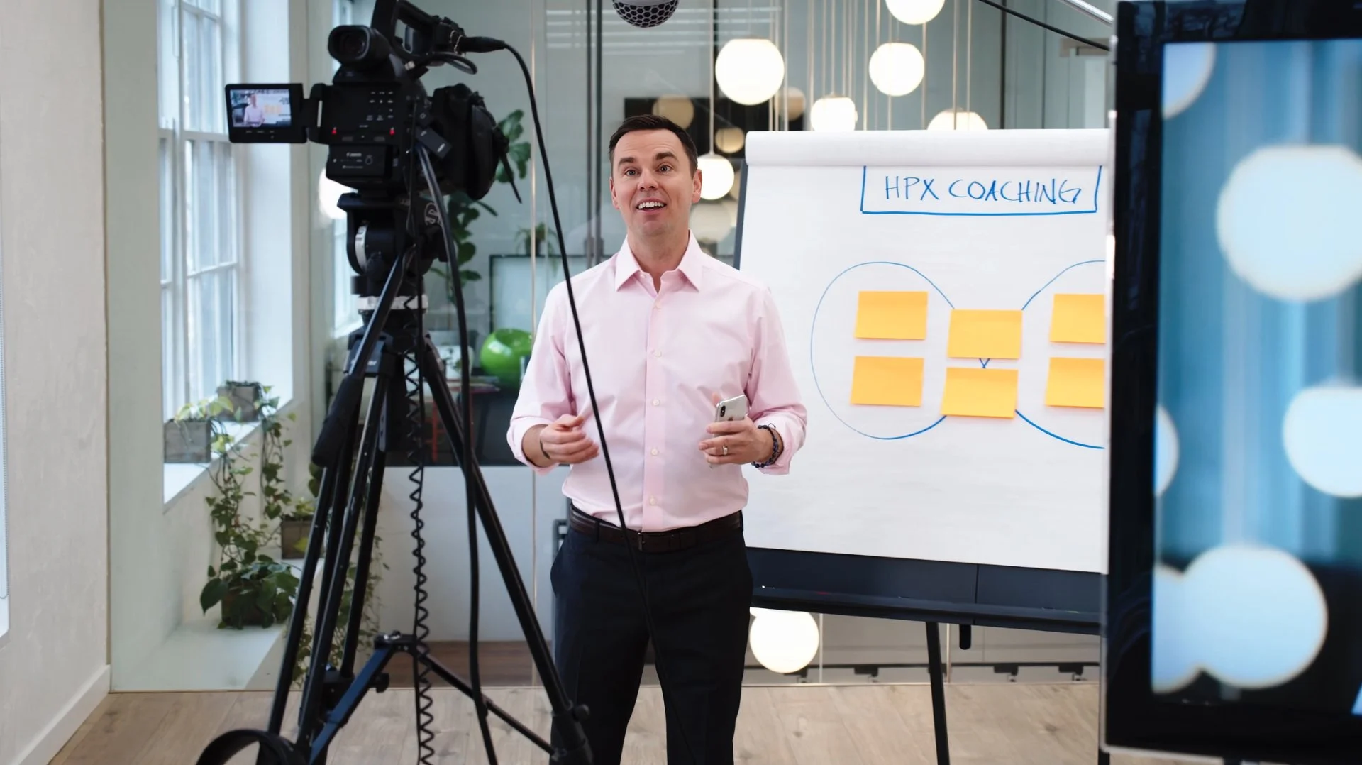 A man in a pink shirt is standing in front of a whiteboard with sticky notes and a diagram labeled 'HPX Coaching'. He is smiling and holding a phone, while being filmed by a professional video camera in a modern office or studio with large windows and decorative lighting.