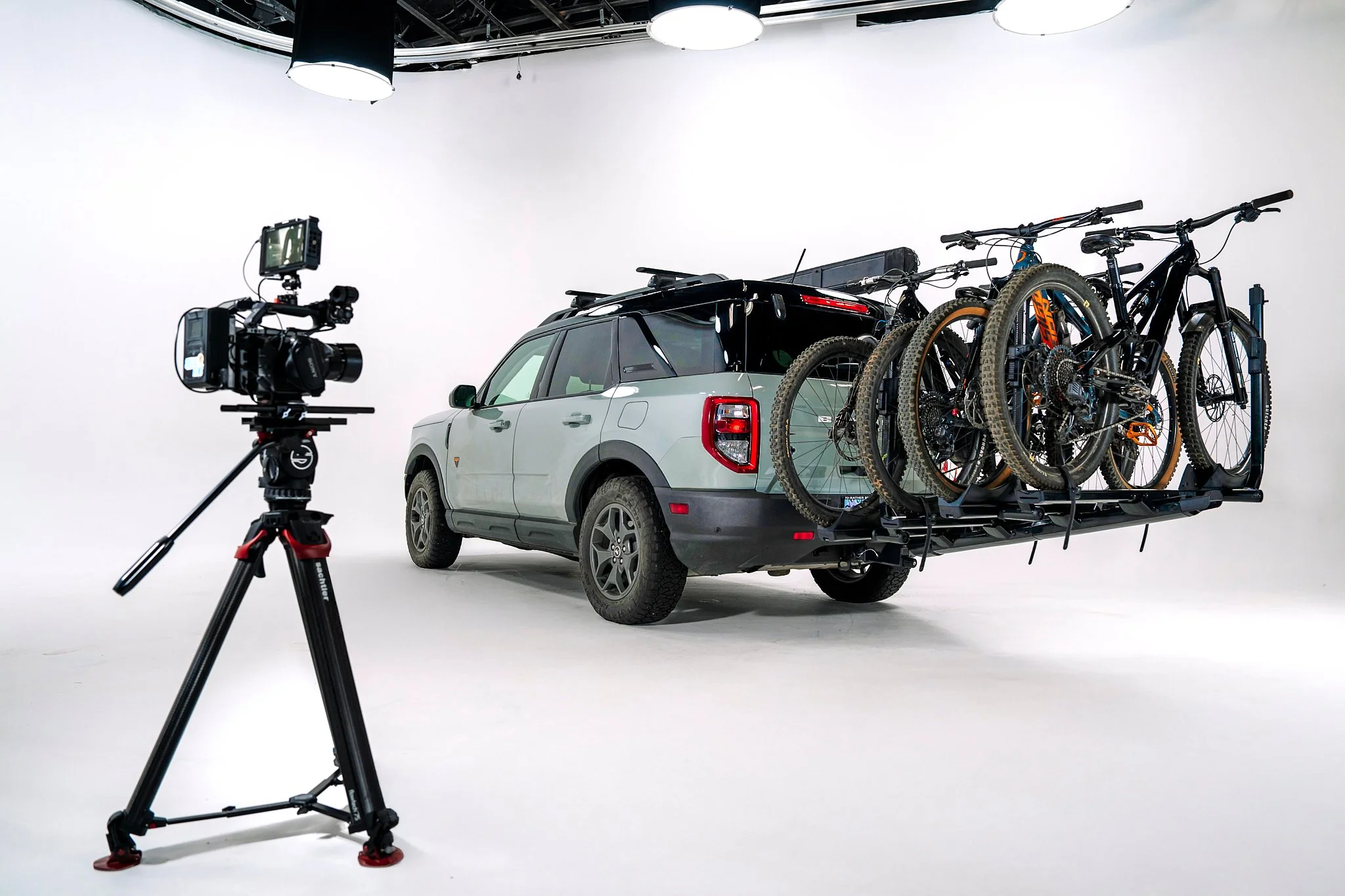 A vehicle transporter with a car and four bicycles on a white background studio setup, with a camera on a tripod pointed at it.