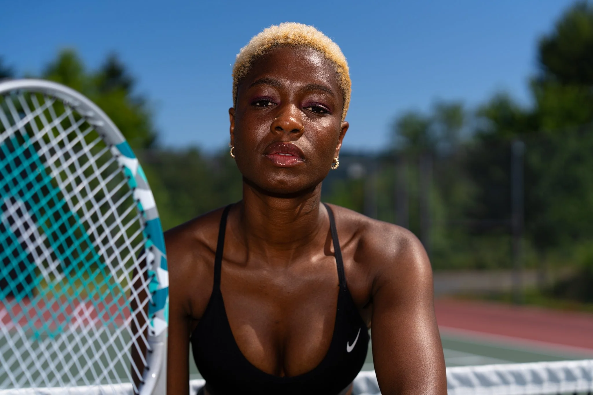 A woman with short blonde hair and dark skin holding a tennis racket outdoors on a sunny day.