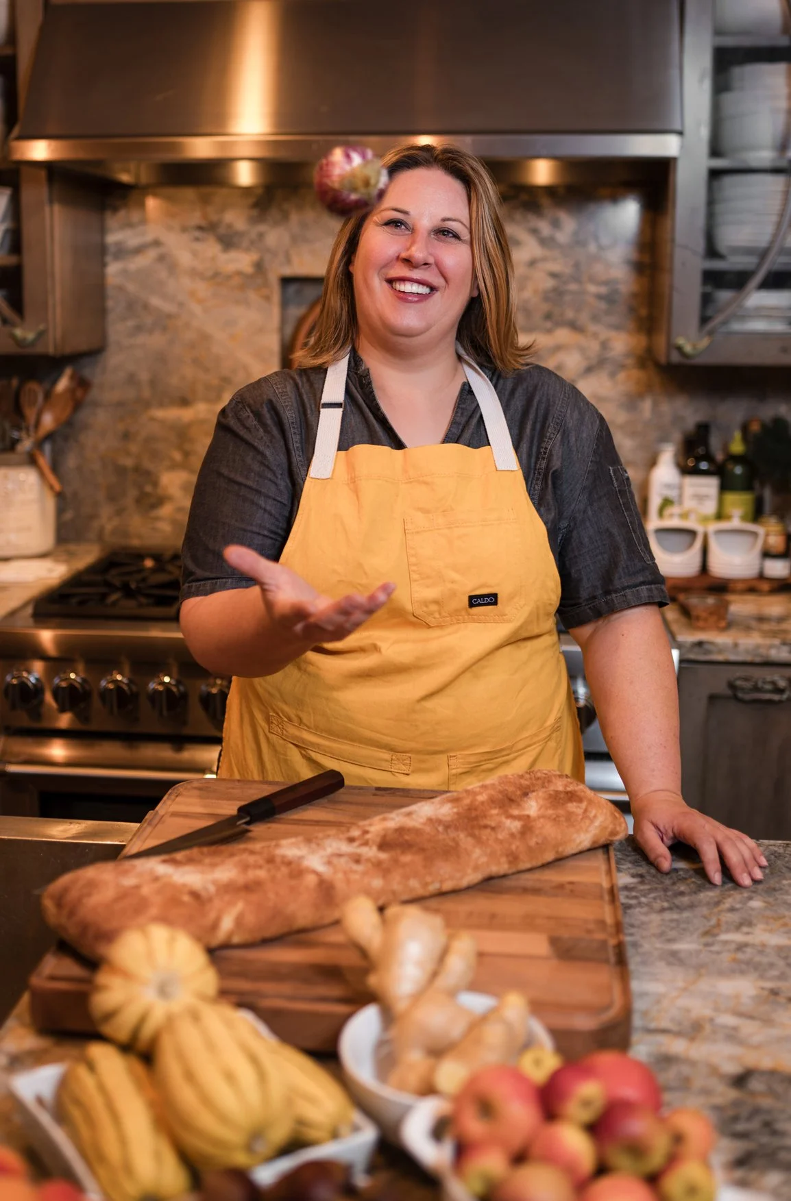 A woman standing in a kitchen with a yellow apron, smiling, and gesturing with her hand. In front of her on a wooden table is a large loaf of bread and a knife. The kitchen has a stone backsplash and various kitchen tools and ingredients on the count