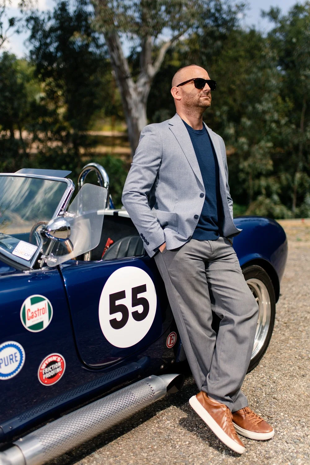 A man in sunglasses, a gray blazer, and gray pants leaning against a vintage blue race car with the number 55 and sponsor decals on its side, outdoors with trees in the background.