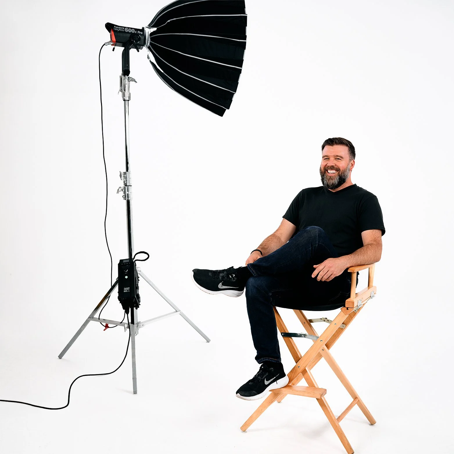 Travis Shields, a man with a beard, wearing a black t-shirt, jeans, and black sneakers, sitting on a wooden director's chair with one leg crossed over the other, smiling in a photography studio. An overhead light is set up nearby.