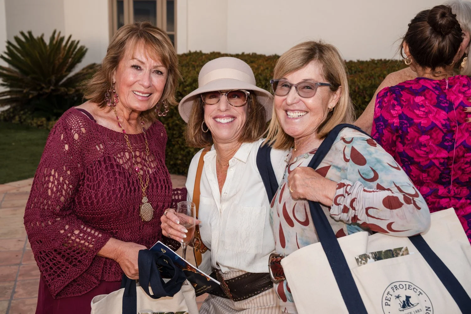 Three women smiling at an outdoor gathering, holding beverages and tote bags, with a bush and part of a white building in the background.