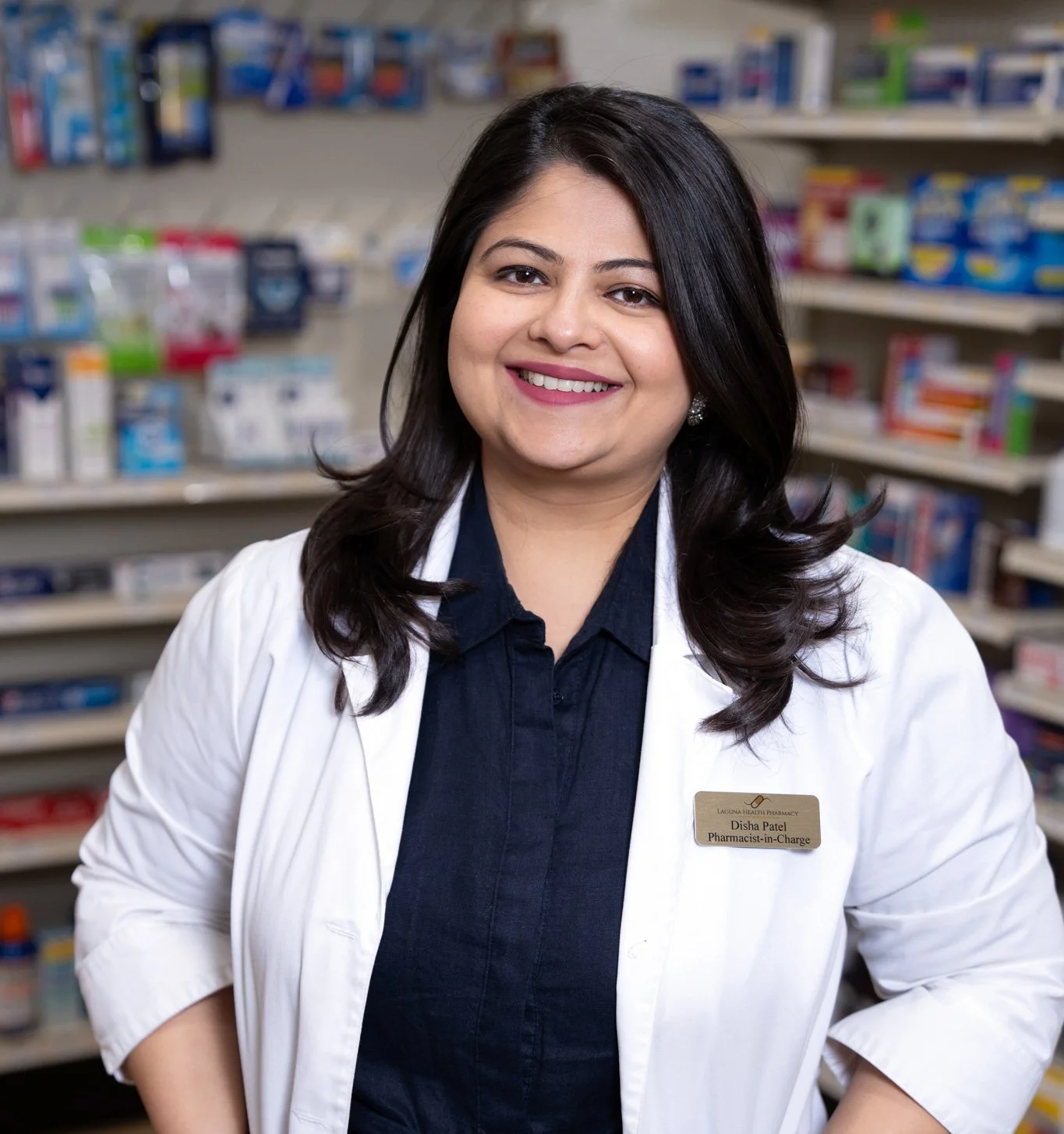 A smiling woman with dark hair and earrings, wearing a white pharmacy coat and a name tag that reads 'Disha Patel, Pharmacist-in-Charge,' standing in front of pharmacy shelves filled with medication bottles and boxes.