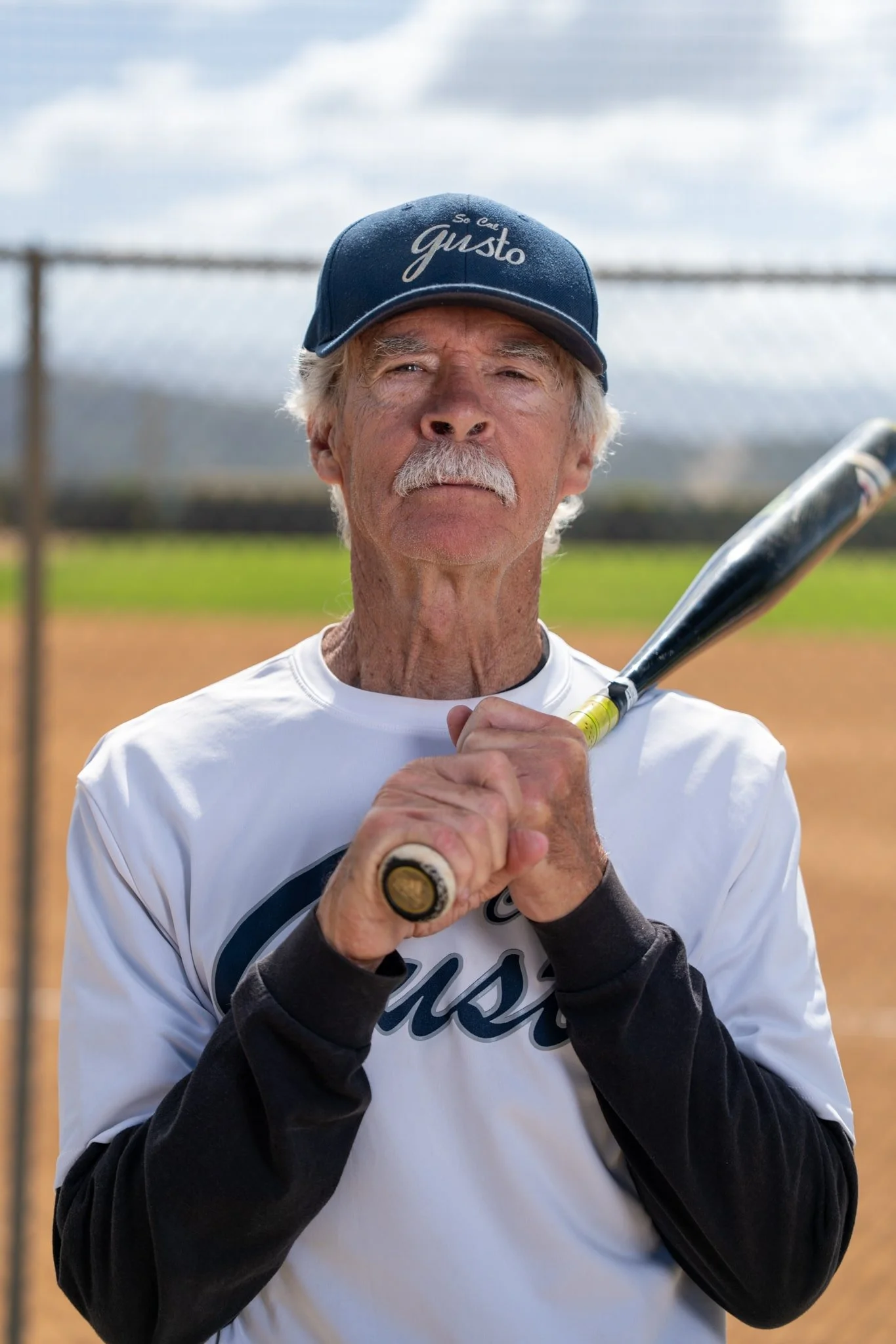 Older man with white hair and mustache holding a baseball bat, wearing a blue cap with the words 'So Gusto', on a baseball field.