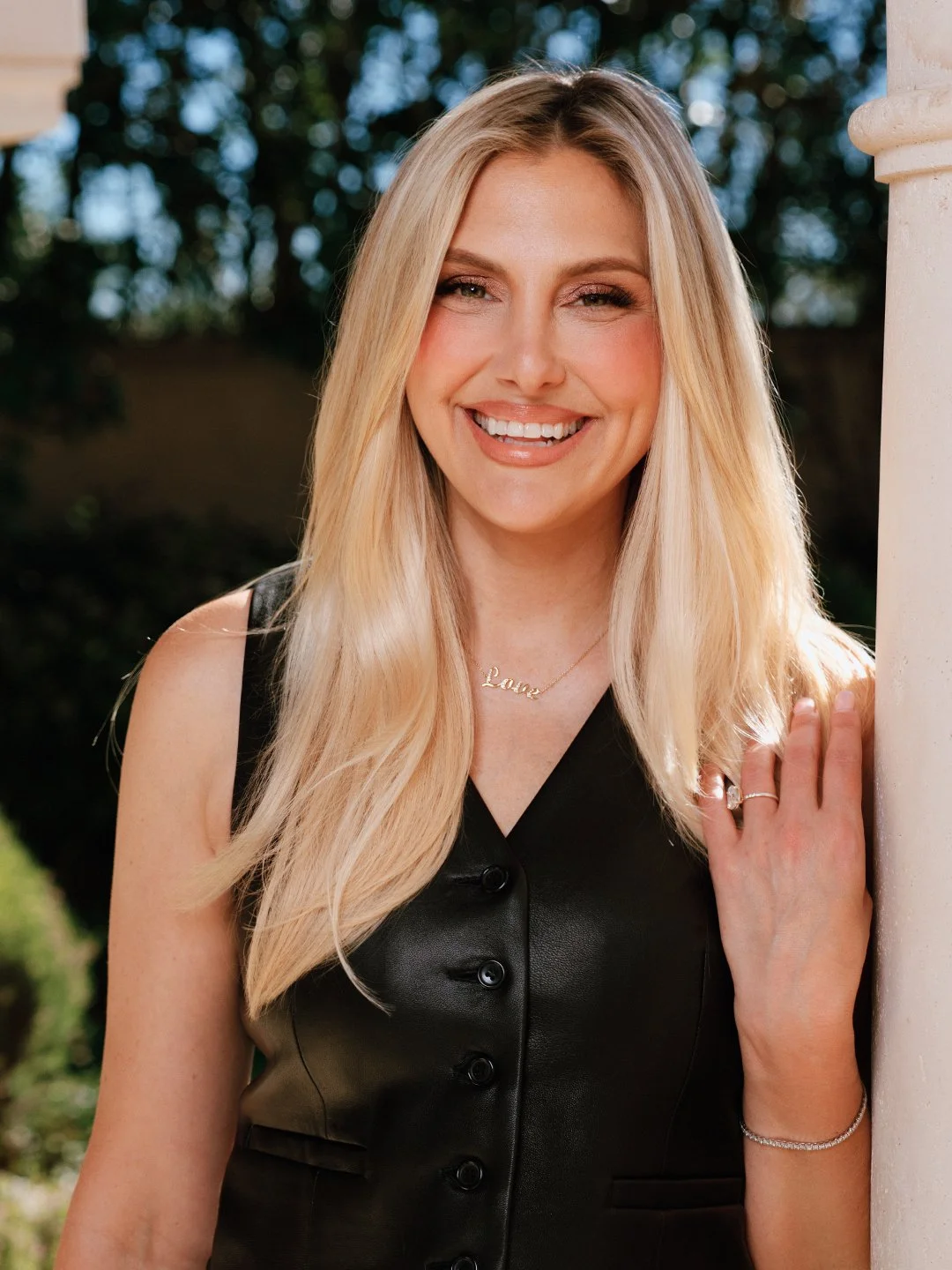 A smiling woman with long blonde hair, wearing a black sleeveless top, a necklace with the word 'Love', and jewelry on her hand and wrist, standing outdoors next to a column.