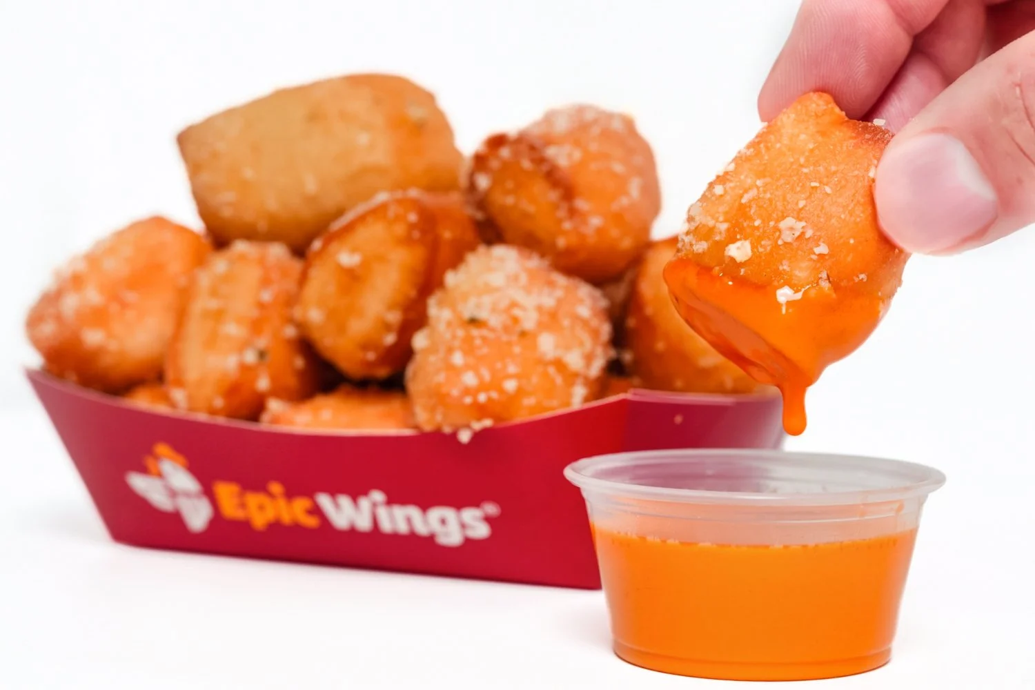 A person dipping a spicy orange-colored chicken nugget into a small container of orange sauce with a food box labeled 'Epic Wings' filled with fried chicken nuggets in the background.