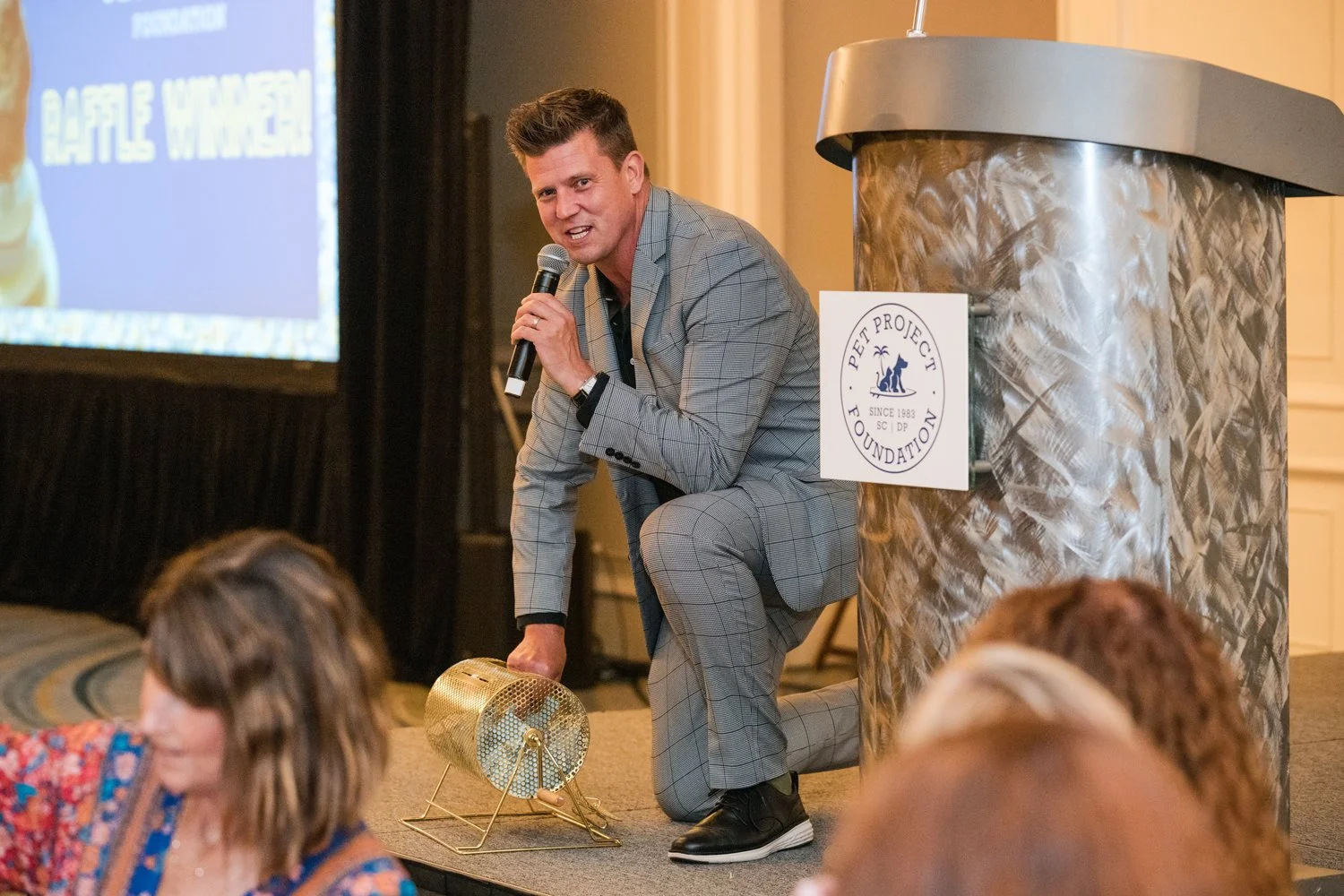 A man in a gray plaid suit crouches on stage, holding a microphone, speaking to an audience, with a sign for Pet Project Foundation on the stage