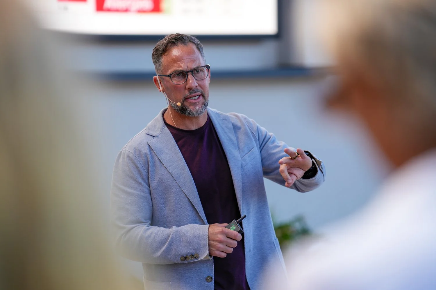 A man with glasses and a beard, wearing a light gray blazer and a dark shirt, is speaking during a presentation or lecture. He is gesturing with his right hand and holding a microphone or remote in his left hand.