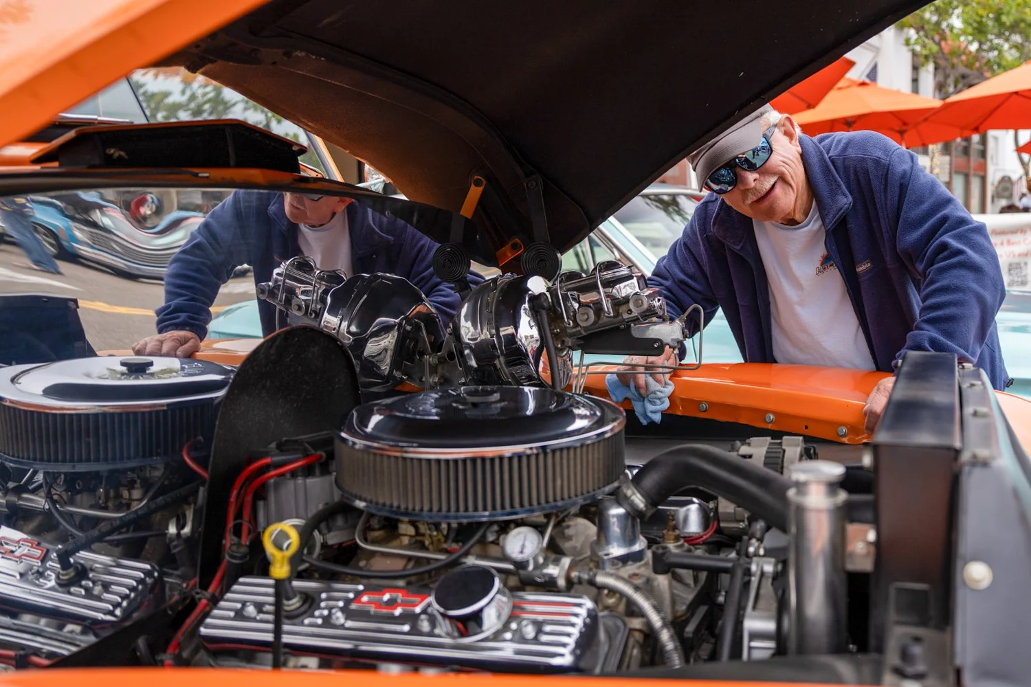 An elderly man with sunglasses and a gray cap, wearing a blue jacket and white t-shirt, working on a car engine at an outdoor car show. The car has an orange hood, and there are orange umbrellas in the background.