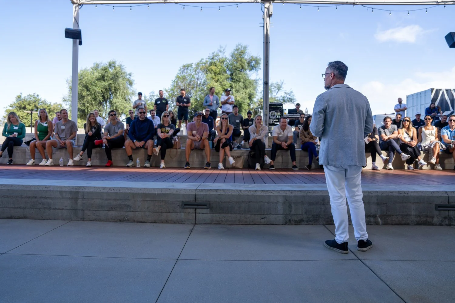 Man in gray blazer speaking outdoors to an audience seated on tiered stone benches, with trees and blue sky in background.