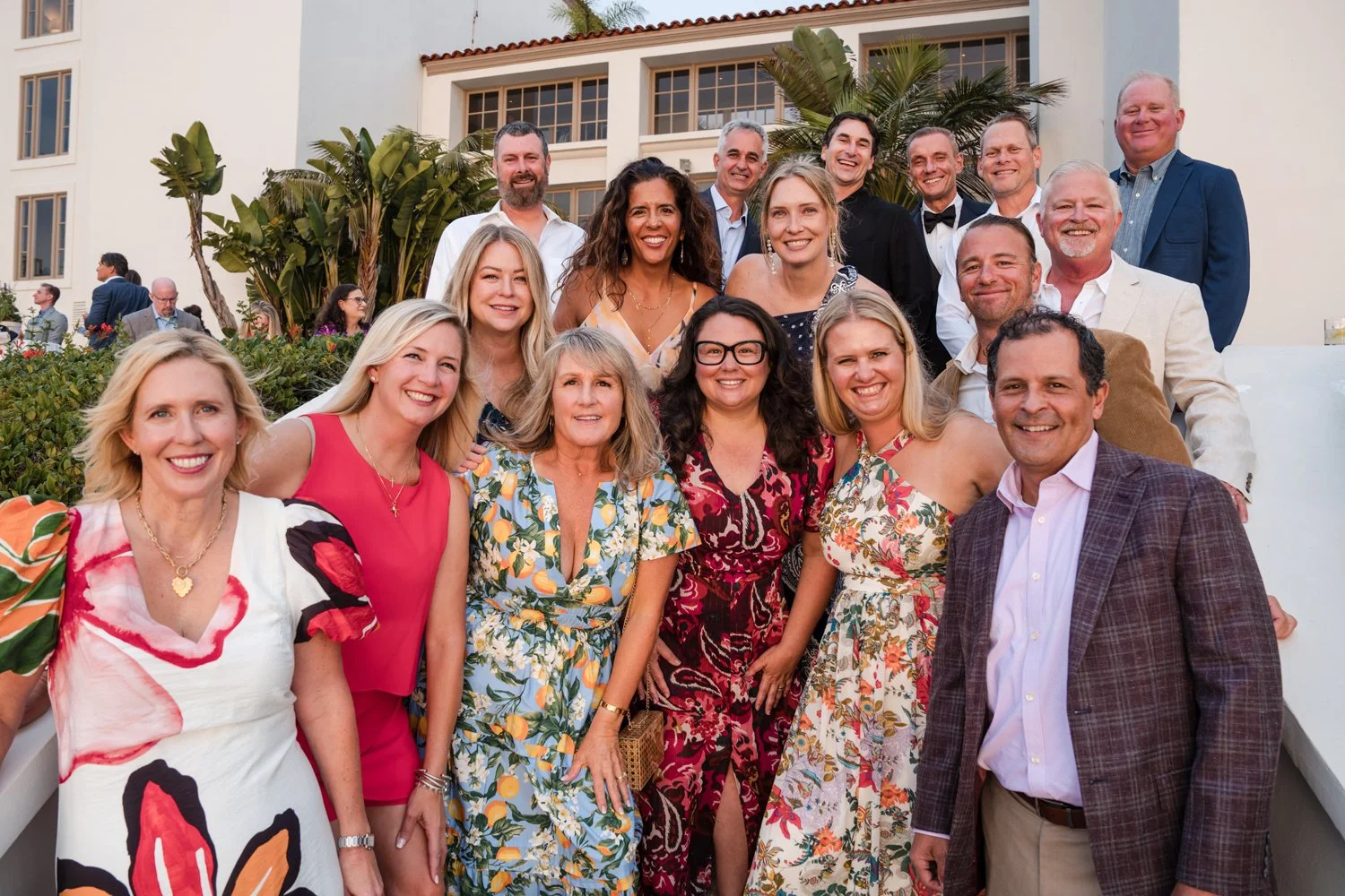 Group of smiling adults at a social gathering outside a white building, with some dressed in colorful outfits and others in formal attire.