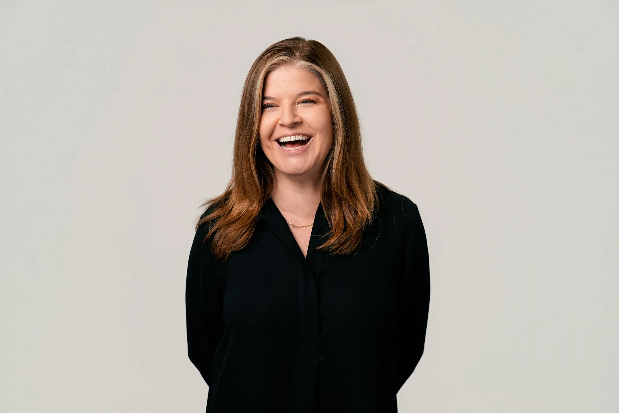 A woman with long, wavy brown hair laughing, wearing a black shirt and a delicate necklace, standing against a plain light gray background.