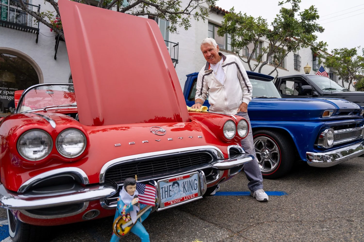 A man is polishing a red vintage Chevrolet Corvette with its hood open, parked next to a blue classic car at a car show on a street lined with trees and white buildings.