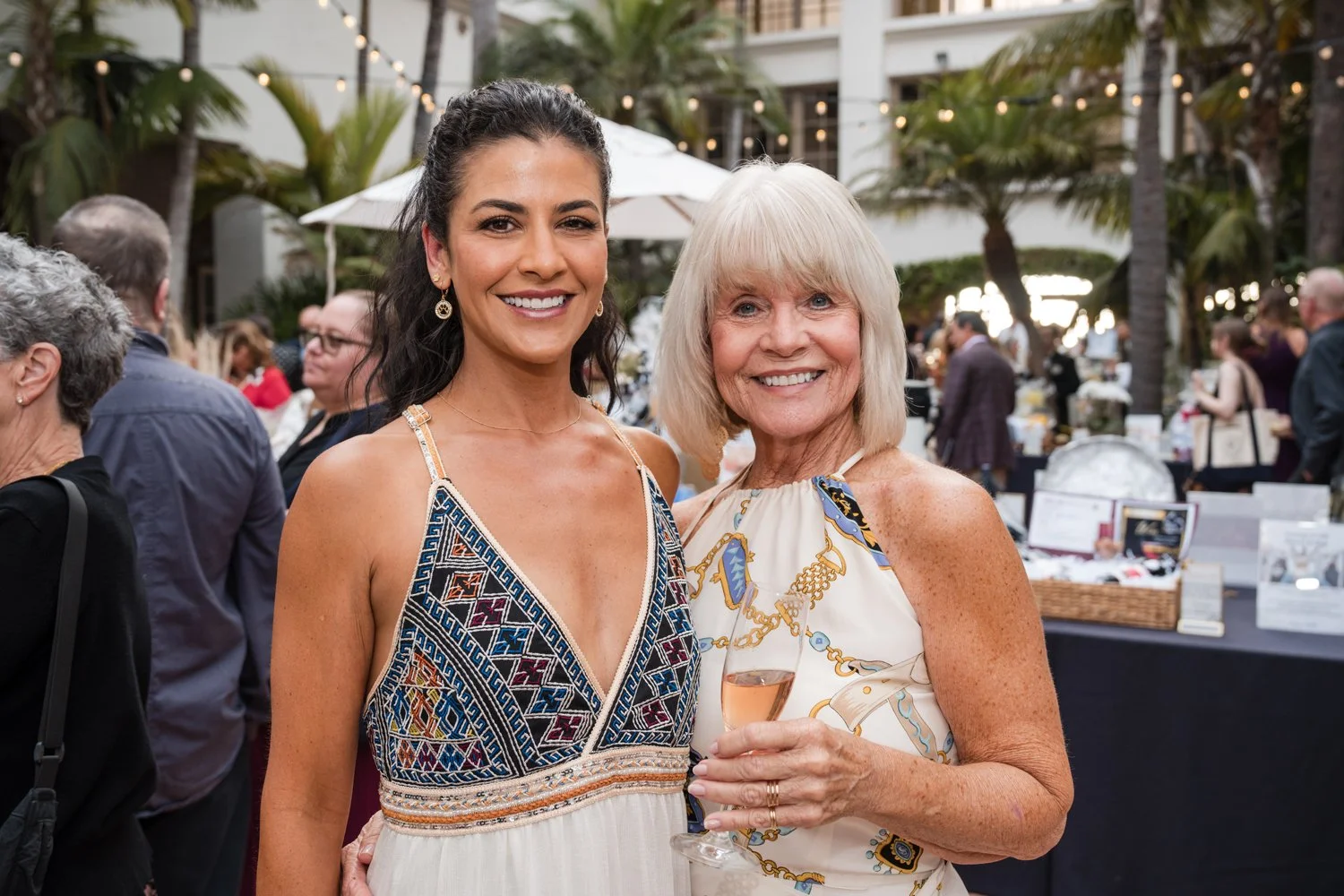 Two women smiling at an outdoor social event, one holding a glass of sparkling wine, surrounded by other guests and string lights in a tropical setting.