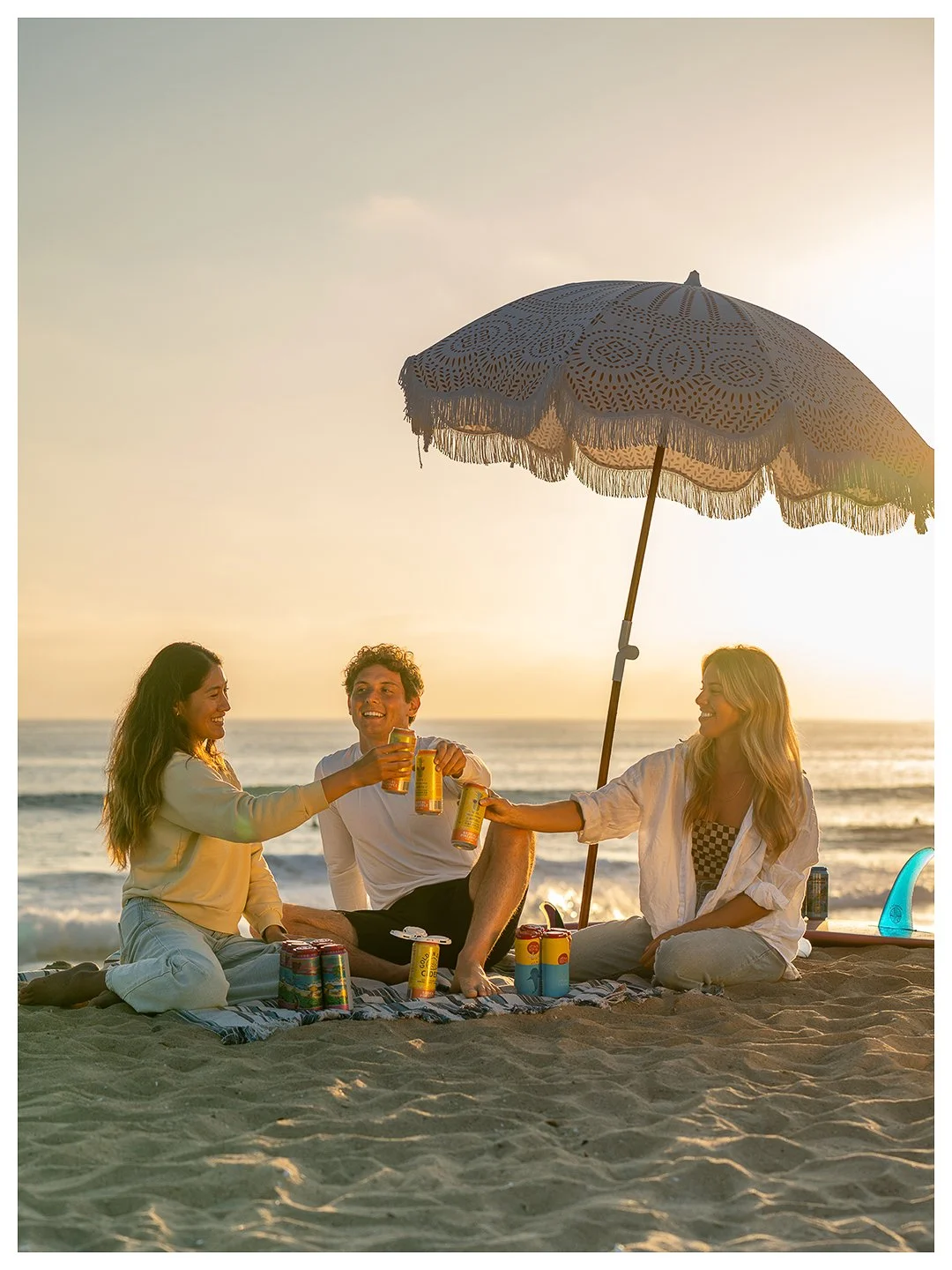 Three friends sitting on the beach under an umbrella, enjoying drinks during sunset.