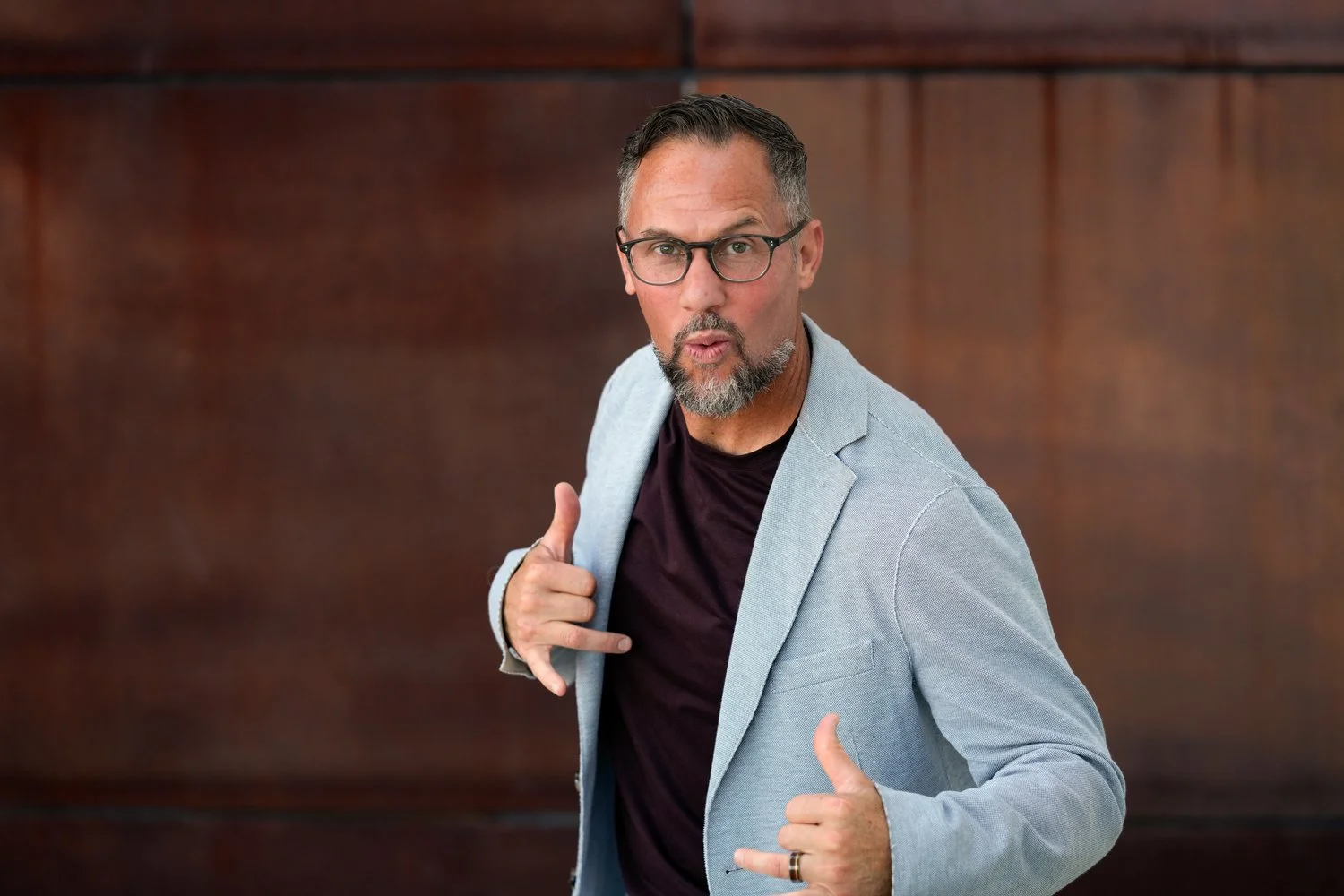 A middle-aged man with glasses, a beard, and gray hair posing with a playful expression, wearing a light gray blazer over a dark shirt, and showing a thumbs-up and shushing gesture.