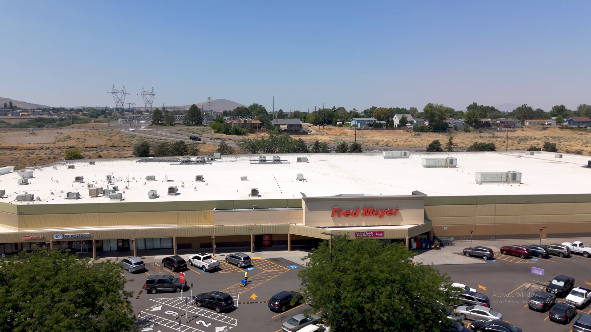 View of a commercial shopping center parking lot with cars, and a large retail store named Fred Meyer on the roof. In the background, a dry landscape with trees, houses, and power lines under a clear blue sky.