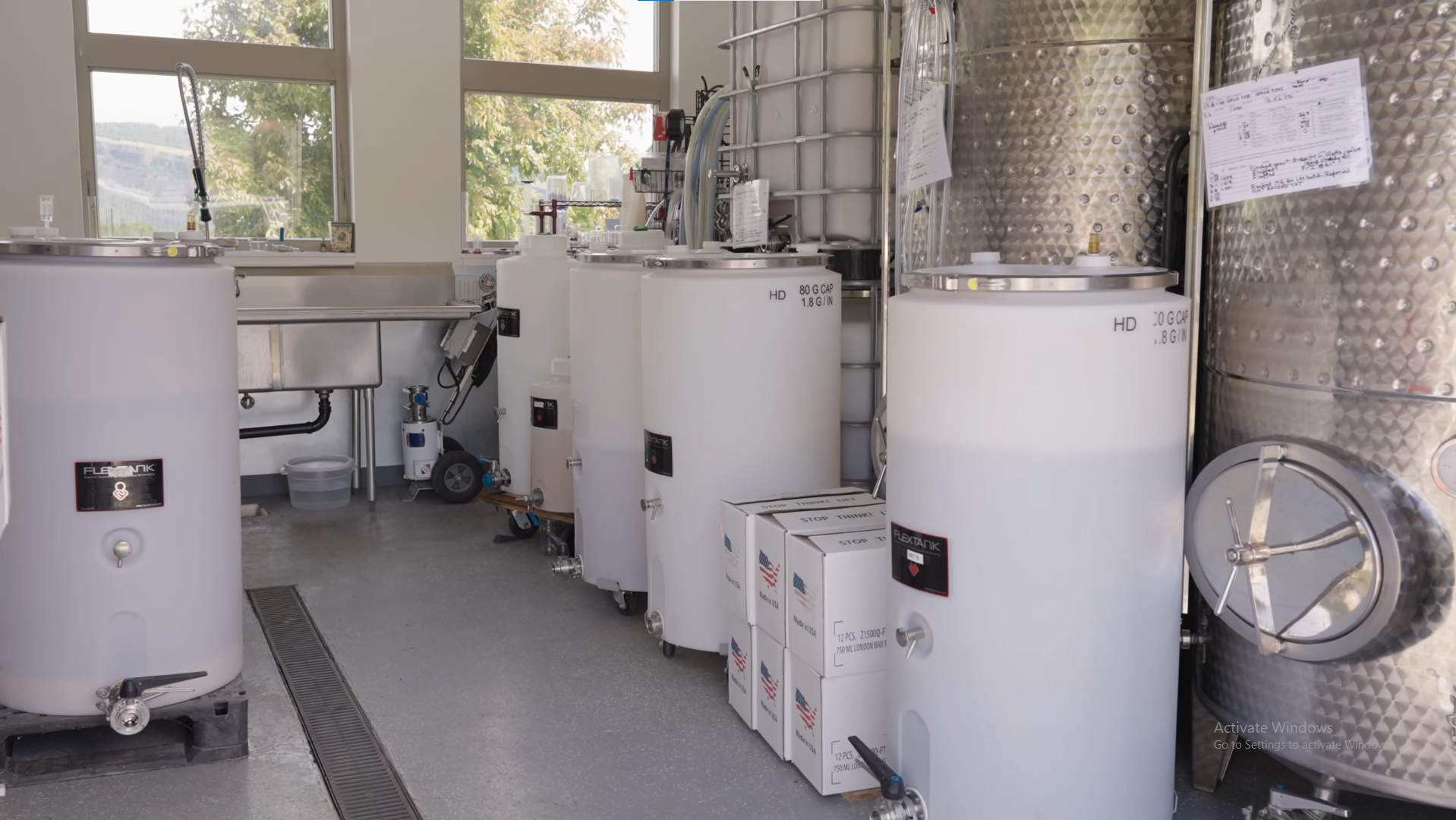 Interior of a laboratory or industrial facility with multiple large white tanks, stainless steel equipment, and boxes labeled as containing laboratory supplies, with windows showing trees outside.