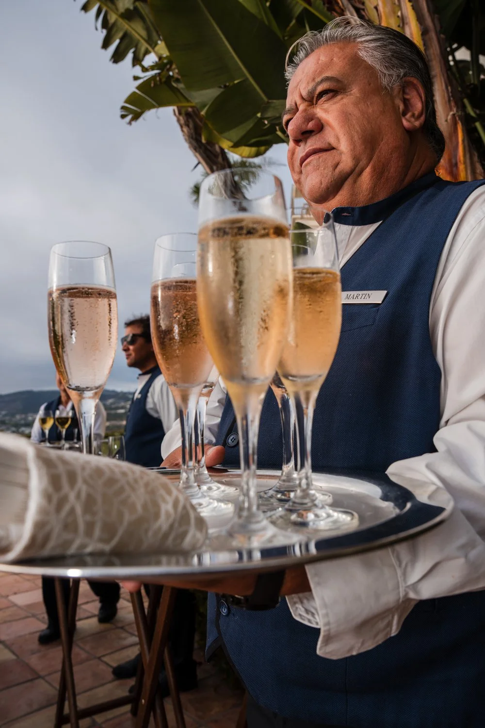 Server holding a tray with four glasses of champagne, with an outdoor setting including palm trees and cloudy sky in the background.