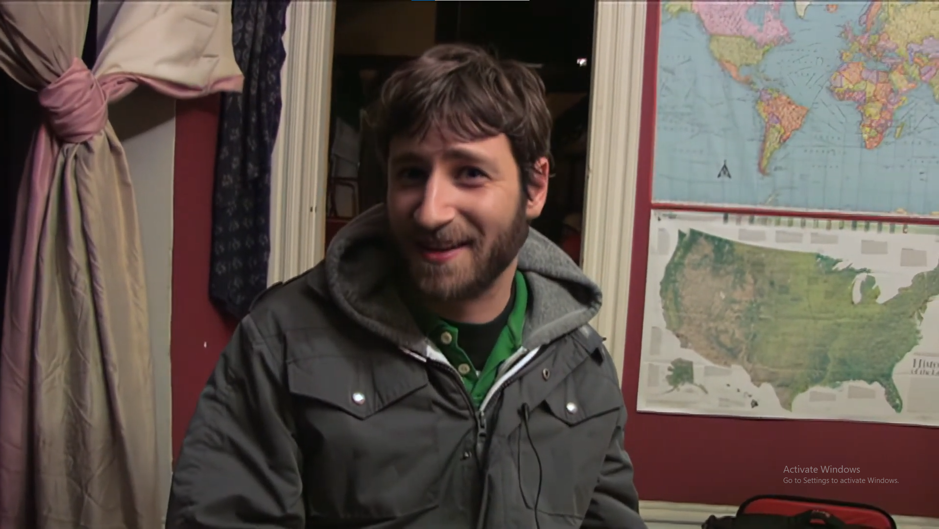 A man with brown hair and a beard smiling, wearing a gray jacket and green shirt, standing in front of world maps on a wall.